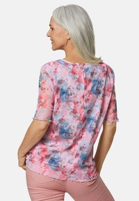 Blumenbluse aus durchsichtigem pinkem Stoff mit blauen und roten Mustern, rundem Halsausschnitt, kurzen Ärmeln mit gewellten Kanten und Reißverschluss auf der Rückseite.