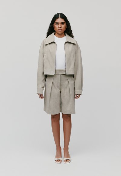 Geknipte beige suède jack met een rits, gecombineerd met bijpassende hooggesneden geplooide shorts. Model draagt een witte top en open teen hakken.