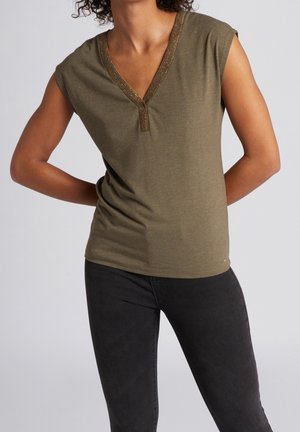 Kvinde iført olivengrøn ærmeløs V-hals top med metallisk kant og sorte bukser, stående med armene bag ryggen mod en grå baggrund.