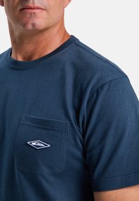 Navyblaues Baumwoll-T-Shirt mit rundem Halsausschnitt, kurzen Ärmeln und einer Brusttasche mit einem weißen gestickten Logo. Glatte Textur.