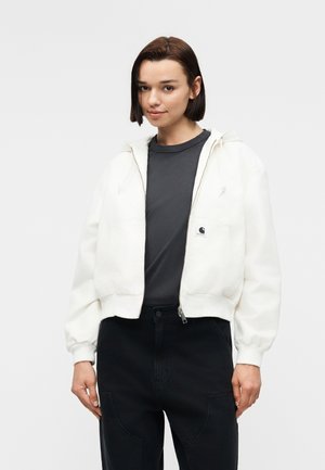 Ung kvinde med kort mørkt hår iført en hvid lynlåshættetrøje, mørkegrå t-shirt og sorte bukser, stående mod en ensfarvet baggrund.