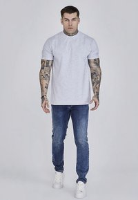 Siva pamučna majica s okruglim izrezom, uparena s plavim trapericama i bijelim tenisicama. Tetovaže su vidljive na rukama. Jednostavan, čist dizajn.