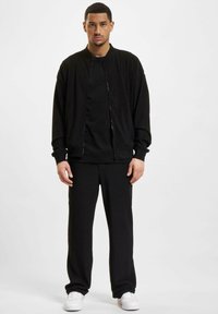 Sort zip-up bomberjakke med lange ærmer, parret med en matchende sort T-shirt og løstsiddende sorte bukser. Hvide sneakers.
