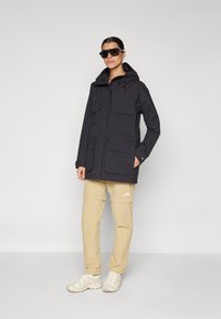 Zwarte waterdichte jas met capuchon, meerdere voorkeuzen, verstelbare koordjes, gecombineerd met beige cargobroek en lichte schoenen.