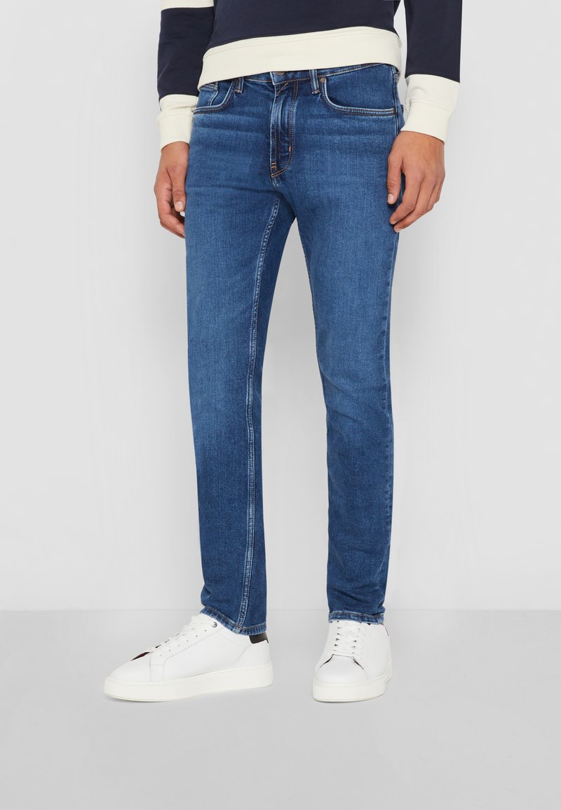 Jean en denim bleu avec une coupe slim, présentant une taille classique et un design à cinq poches. Portés avec des baskets blanches, affichant un look décontracté.