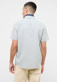 Koszula z krótkim rękawem w jasnoszarym kolorze, wykonana z tkaniny o subtelnej teksturze, z granatowym wzorzystym kołnierzykiem i zaokrąglonym dołem.