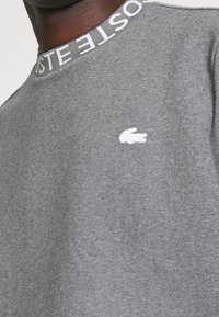 Grå sweatshirt med rund hals och en vit krokodil-logga på bröstet samt "LACOSTE" text på kragen.