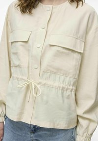 Béžová skrátená bunda vyrobená z ľahkého materiálu s dvoma prednými vreckami, sťahovacím pásom a elastickými rukávmi. Hladká textúra.