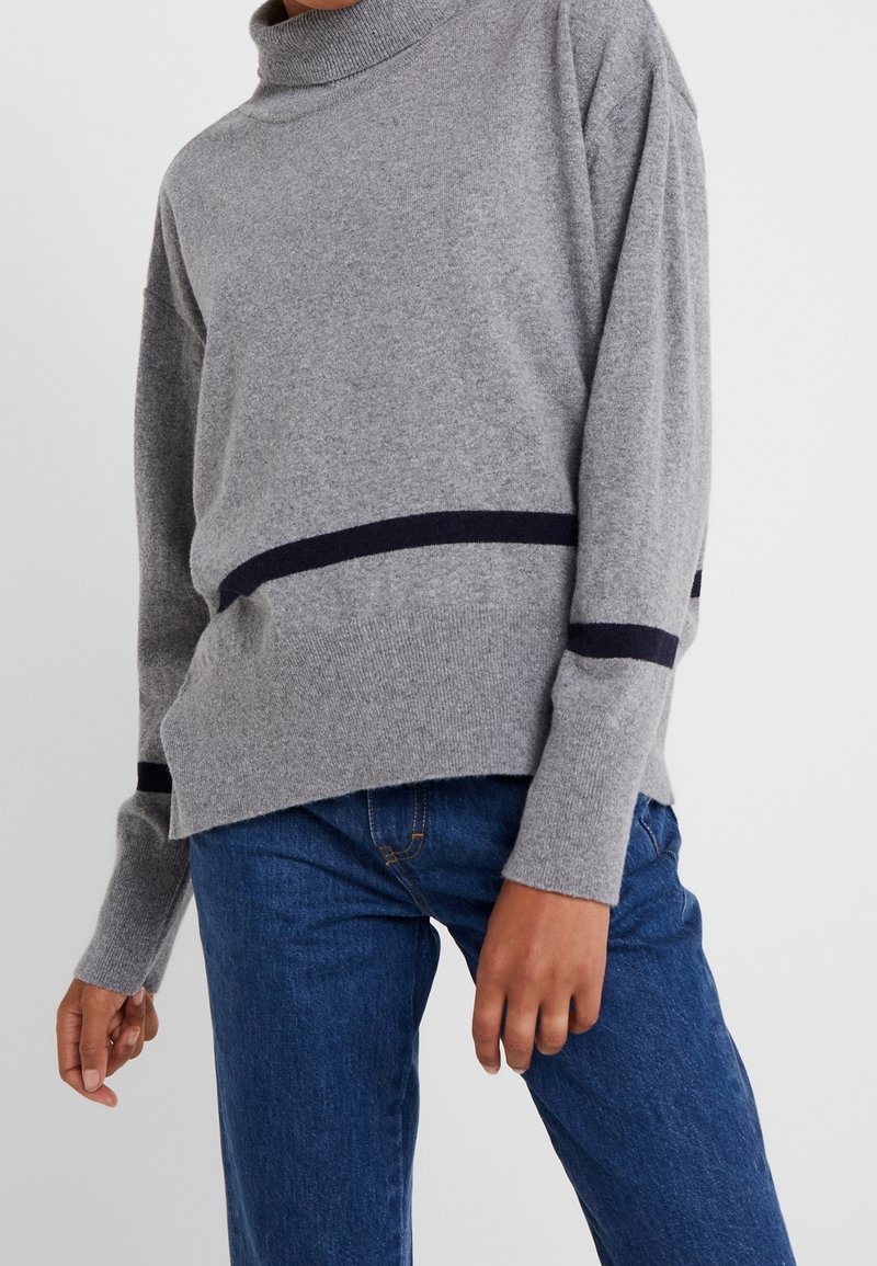 Pull à col roulé en maille grise avec des rayures bleu marine à l'ourlet et aux manches, caractérisé par une coupe décontractée et une texture douce, associé à un jean en denim bleu.