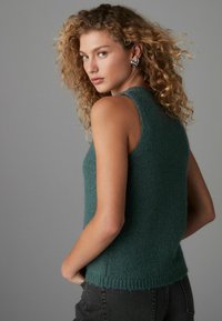 Dunkelgrüne ärmellose Strickweste mit weicher Textur, geripptem Saum und Rundhalsausschnitt. Kombiniert mit schwarzen Jeans.