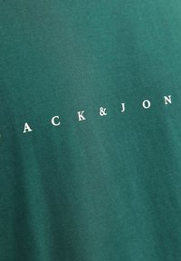 Grünes Baumwoll-T-Shirt mit dem Aufdruck "ACK & JON" in weißen Buchstaben. Weiche Textur, klassischer Rundhalsausschnitt, minimalistischer Stil.