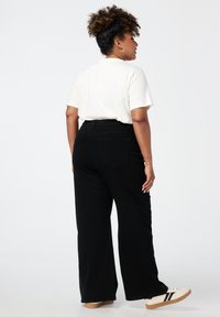 Pantalon large noir à taille haute et poches arrière, associé à une chemise blanche à manches courtes et des baskets beiges. Texture de tissu douce.