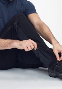 Mann trägt schwarze Hose und Schuhe, sitzt und zieht einen Reißverschluss am unteren Bein der Hose hoch.