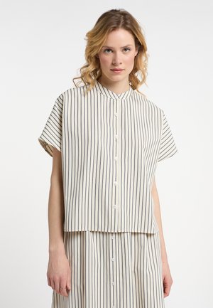 Femme aux cheveux clairs et ondulés portant une chemise blanche à manches courtes boutonnée avec de fines rayures noires verticales et une jupe rayée assortie.