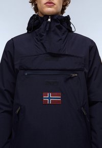 Jeune personne portant une veste à capuche bleu marine avec une poche zippée à l'avant et un patch du drapeau norvégien sur la poitrine, sur un fond uni.