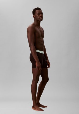 Homme mince à la peau foncée, debout pieds nus en boxer noir avec une ceinture blanche sur fond gris uni.