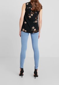 Žena v černém bezrukávovém květinovém topu, světle modrých slim džínech a černých vysokých podpatcích, stojící na jednobarevném pozadí.