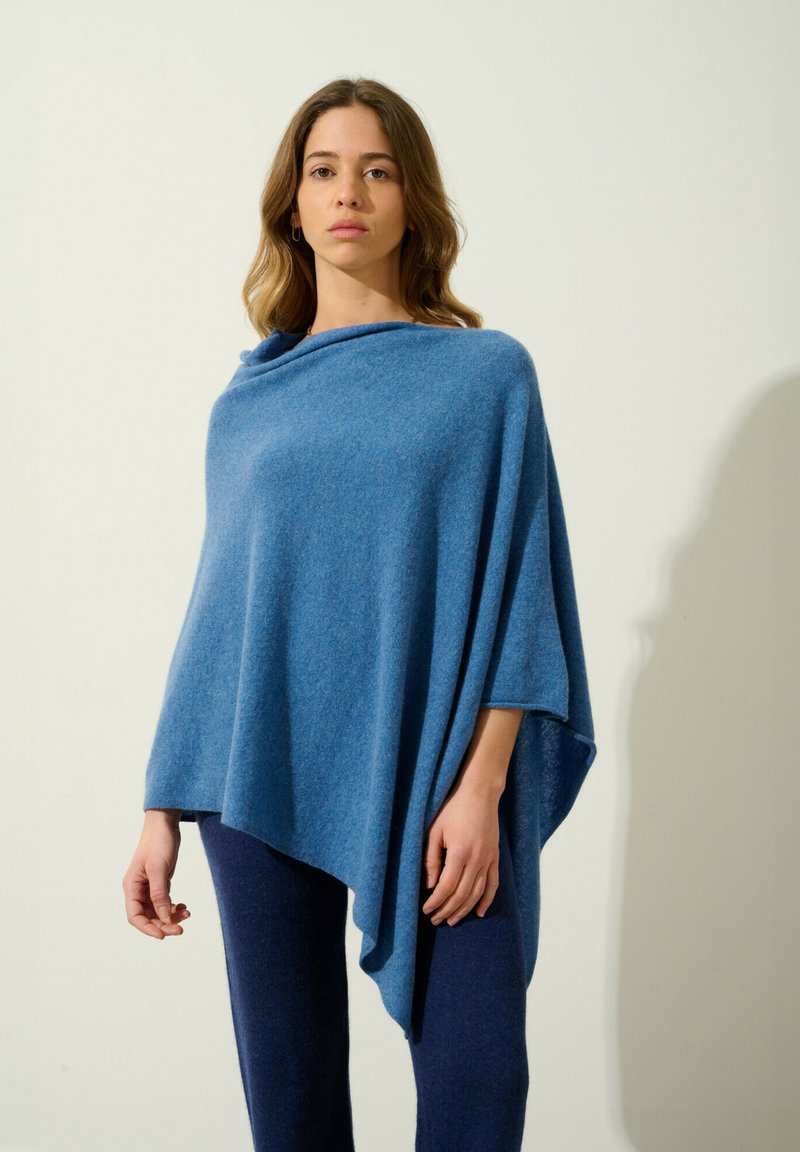Blå ullponcho med draperad design, som har en rund halsringning och asymmetrisk nederdel, kompletterad med mörkblå tajta byxor.