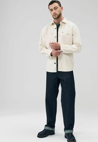 Weißes Hemd mit Knopfleiste und zwei Brusttaschen, kombiniert mit dunkelblauen Jeans und schwarzen Stiefeln mit umgeschlagenem Saum. Einfaches, strukturiertes Design.