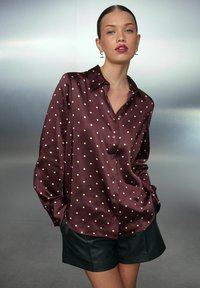 Mujer vistiendo una blusa de satén burdeos con lunares, combinada con pantalones cortos de cuero negros, de pie con las manos en los bolsillos frente a un fondo neutro.