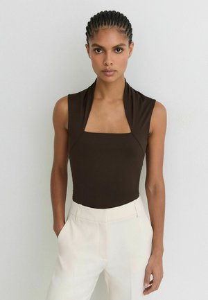 Femme aux cheveux tressés portant un haut marron foncé sans manches à col carré et un pantalon écru taille haute, debout contre un mur uni.
