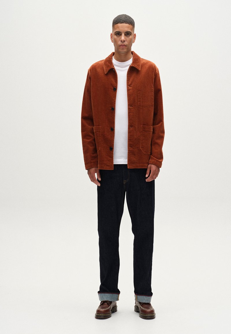 En bränd orange kordfloretjacka med krage, framknappar och två fickor; matchad med en vit t-shirt och mörka jeans. Bruna skor.
