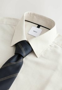 Chemise beige à boutons avec un tissage texturé et une étiquette noire ; associée à une cravate bleu foncé à rayures diagonales et accents beiges.