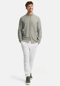 Camisa abotonada gris clara con un patrón texturizado, combinada con pantalones de lino blancos y zapatillas gris; de pie en un fondo neutro.