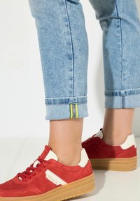 Sneakers in suede rosso con dettagli crema, caratterizzate da un design sportivo e una suola spessa in gomma colorata. Pantaloni di denim azzurro chiaro arrotolati con un'etichetta gialla.