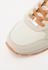 Beige tenisky s texturovanou tkaninou a hladkými akcenty, s hnědými tkaničkami a gumovou podrážkou. Stříbrná logo visačka na boku.