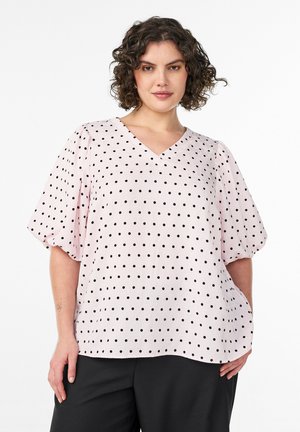 Kvinde med krøllet hår iført en lyserød bluse med sorte polkaprikker og sorte bukser, stående mod en hvid baggrund.