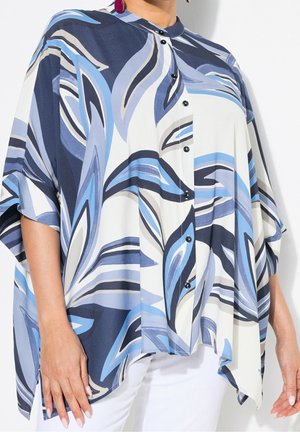Femme portant un chemisier ample boutonné avec des motifs abstraits de feuilles bleues, blanches et noires et un pantalon blanc.