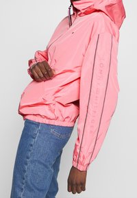 Růžová lehká bunda s kapucí a elastickými manžetami, na které je umístěno logo na rukávu a černé lemování. Návrh k modrým džínům.