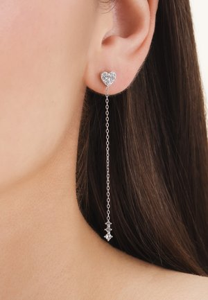 Zilveren oorbel in de vorm van een hartje met kleine kristallen en een lange, delicate ketting die eindigt in een kleine kristallen cluster, gedragen aan het oor van een vrouw.