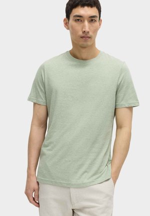 Jonge man die een lichtgroen T-shirt met korte mouwen en beige broek draagt, staand met één hand in zijn zak tegen een effen achtergrond.