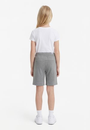 Barn med langt blond hår, der står med ryggen til, iført hvid t-shirt, grå knælange shorts, hvide sokker og hvide sneakers på hvid baggrund.