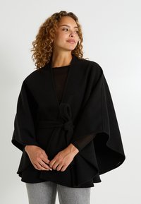 Cape de mezcla de lana negra con mangas anchas, cintura con lazo y forma acampanada. La modelo lleva una blusa negra y pantalones grises.