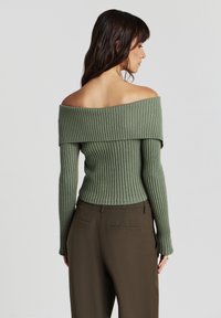 Frau mit dunklem Haar, die einen schulterfreien, gerippten grünen Pullover und braune Hosen trägt, steht seitlich vor einem schlichten hellen Hintergrund.