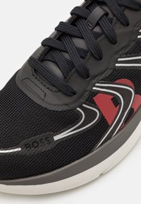 Svart sneakers med överdel av mesh, gummidetaljer och röd och vit dekoration. Har en framträdande logotyp, kontrasterande texturer och plana snörningar.