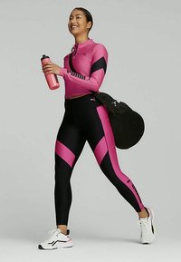 A Puma aktív öltözék szettje egy rózsaszín hosszú ujjú felsőből és fekete leggingsből áll, rózsaszín geometriai elemekkel, kiegészítve egy vízflaskával és egy edzőtáskával.