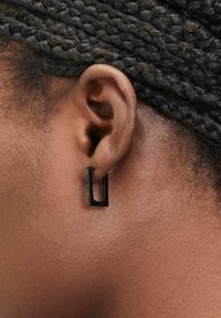 Schwarze rechteckige Hängehänger, mit mattem Finish und minimalistischem Design. Getragen am gepiercten Ohr, das die strukturierte Haut und geflochtenes Haar zeigt.