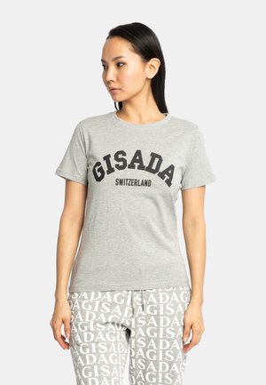 Femme aux longs cheveux foncés porte un T-shirt gris avec le texte "GISADA Switzerland" et un pantalon gris assorti avec un motif répétitif "GISADA".