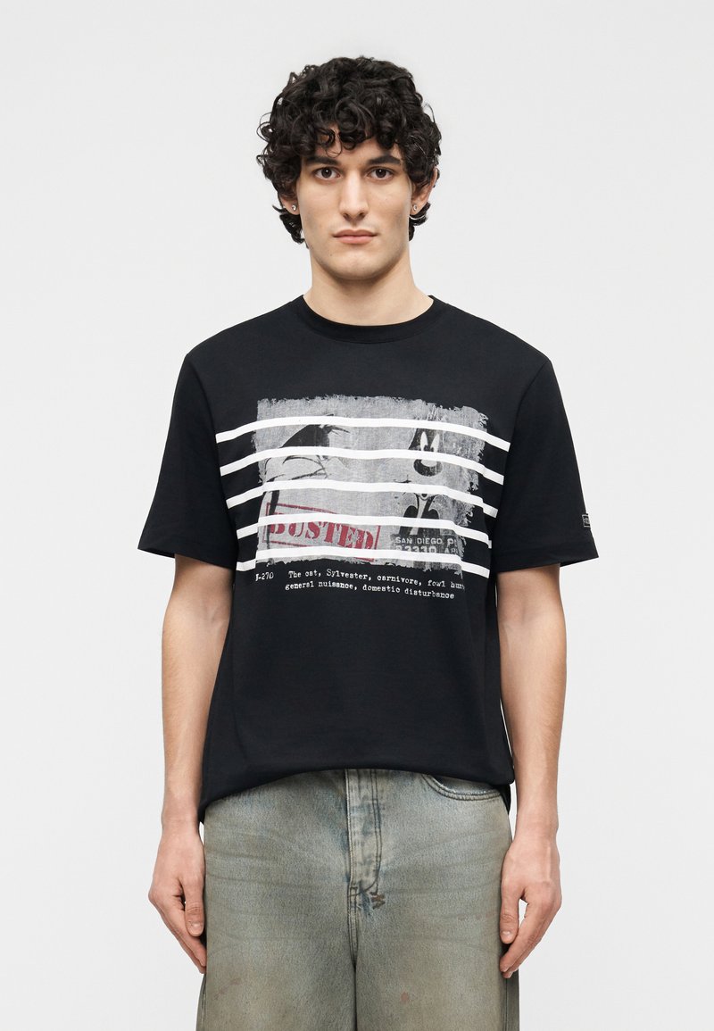Jeune homme aux cheveux bouclés portant un t-shirt noir avec un personnage de dessin animé en noir et blanc derrière des barreaux blancs et un texte en dessous.