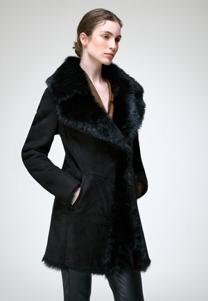 Femme portant un manteau noir doublé de fourrure, mains dans les poches, regardant sur le côté sur un fond clair uni.