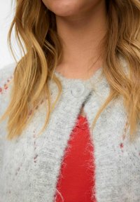 Graue Strickjacke mit weicher Textur, die einen großen Knopf am Hals besitzt und über einem roten Oberteil getragen wird. Sichtbare helle rote Akzente.