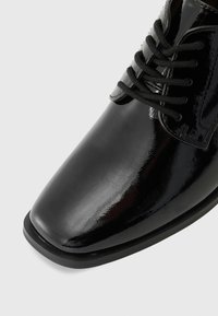 Black patent leather shoe with a rounded toe, smooth surface, and lacing. Features a low, flat black sole.