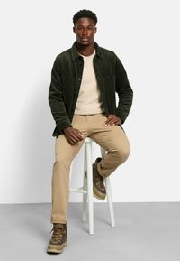 Forest green corduroy shirt jacket, beige sweater, tan pants, and brown hiking boots with thick soles. White stool and gray background.