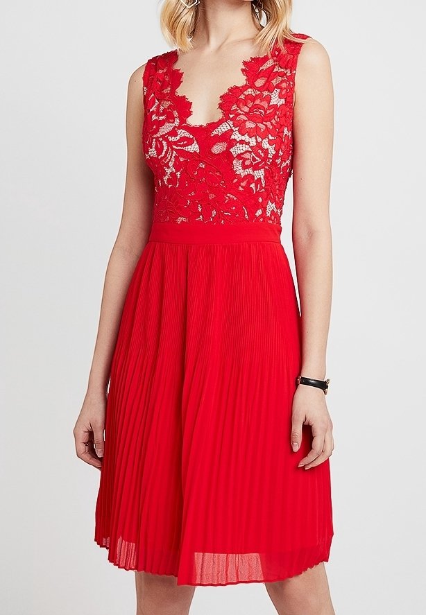 Femme portant une robe rouge sans manches avec un corsage en dentelle florale et une jupe plissée, se tenant devant un fond uni.