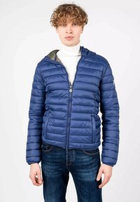 Giacca blu gonfiabile con cuciture orizzontali, chiusura con zip e cappuccio. Abbinata a un dolcevita bianco e jeans blu scuri.