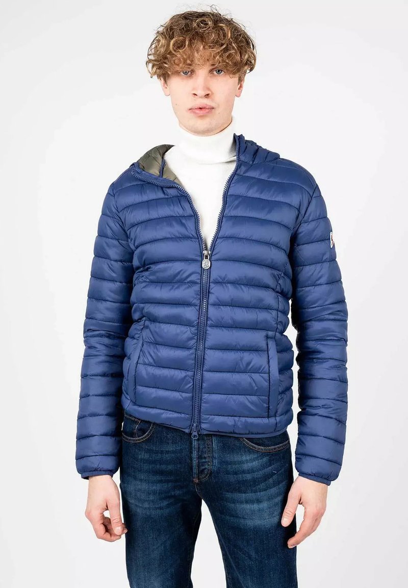 Giacca blu gonfiabile con cuciture orizzontali, chiusura con zip e cappuccio. Abbinata a un dolcevita bianco e jeans blu scuri.